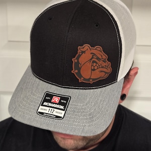 May include: A black and grey trucker hat with a white mesh back. The hat has a brown leather patch with a bulldog design. The hat has a black label with the text "Richardson 112 Trucker Adjustable" on it.