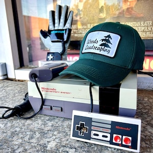 May include: A green baseball cap with a white patch that says "Woods Landscaping" and a small pine tree graphic. The cap is sitting on top of a vintage Nintendo Entertainment System with a controller.