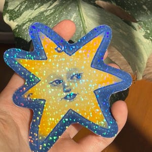 Könnte beinhalten: Ein sternförmiges Ornament mit einem gelben Sonnengesicht, blauen Augen und Lippen, umgeben von einem dunkelblauen Rand. Die gesamte Oberfläche ist mit einem holografischen Glitzer-Finish versehen, das Licht in verschiedenen Farben reflektiert.