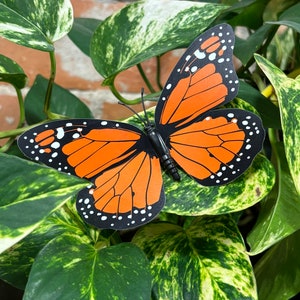May include: A decorative butterfly with orange wings and black and white details, perched on green and yellow variegated leaves. The butterfly has a black body and antennae. The background features a brick wall.