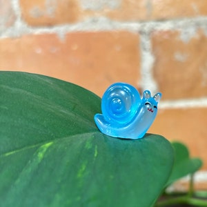 May include: A small, translucent blue snail figurine with a smiling face and a spiral shell. The snail is perched on a large, dark green leaf, with a blurred brick wall in the background. The figurine is a whimsical decorative item.