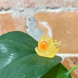 May include: A small, yellow snail figurine with an orange spiral shell and a smiling face. The figurine is perched on a large, dark green leaf with lighter green veins. The background is a brick wall.