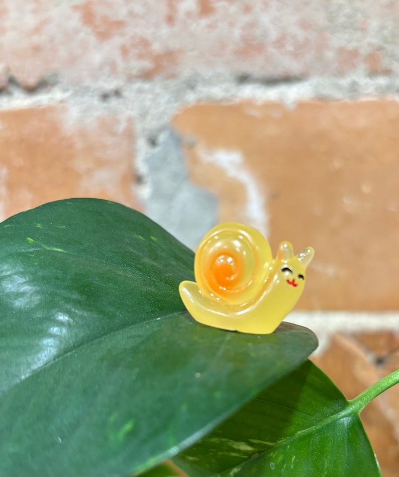 May include: A small, yellow snail figurine with an orange spiral shell and a smiling face. The figurine is perched on a large, dark green leaf with lighter green veins. The background is a brick wall.