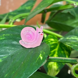 May include: A small, pink, translucent snail figurine with a smiling face and a red mouth. The snail is perched on a large, green leaf, with other green leaves and stems in the background. The figurine has a glossy finish.