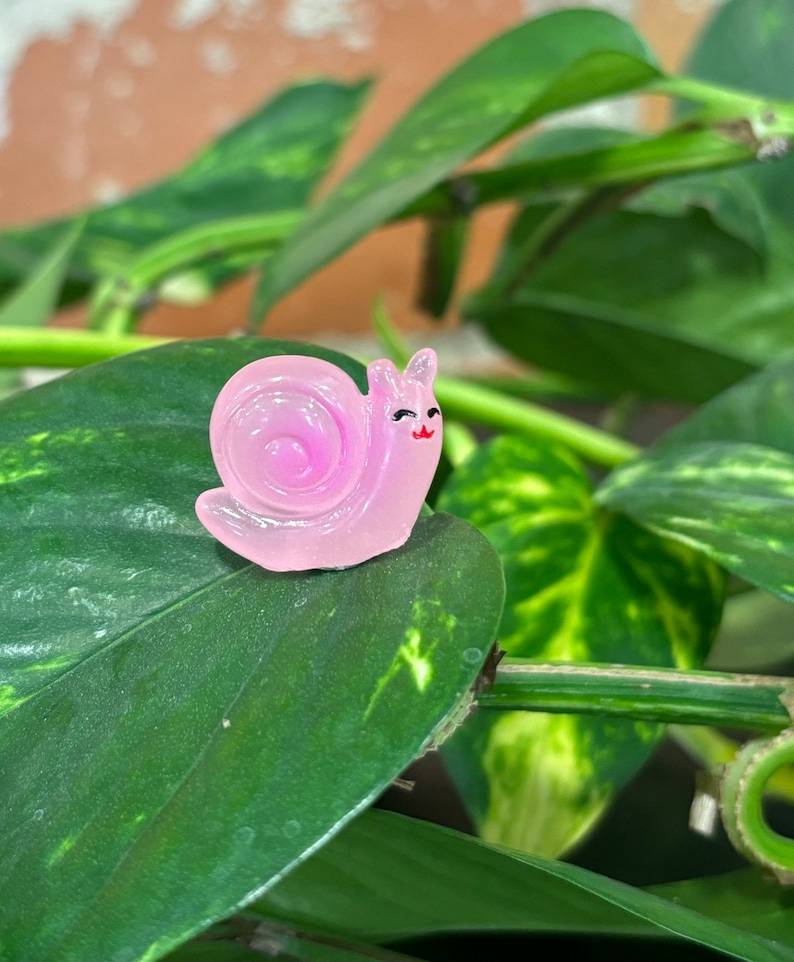May include: A small, pink, translucent snail figurine with a smiling face and a red mouth. The snail is perched on a large, green leaf, with other green leaves and stems in the background. The figurine has a glossy finish.