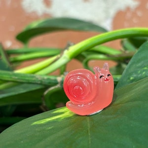 May include: A translucent, coral-colored snail figurine with a smiling face and black eyes. The snail is positioned on a large, green leaf, with a blurred background of more green leaves and a brick wall.