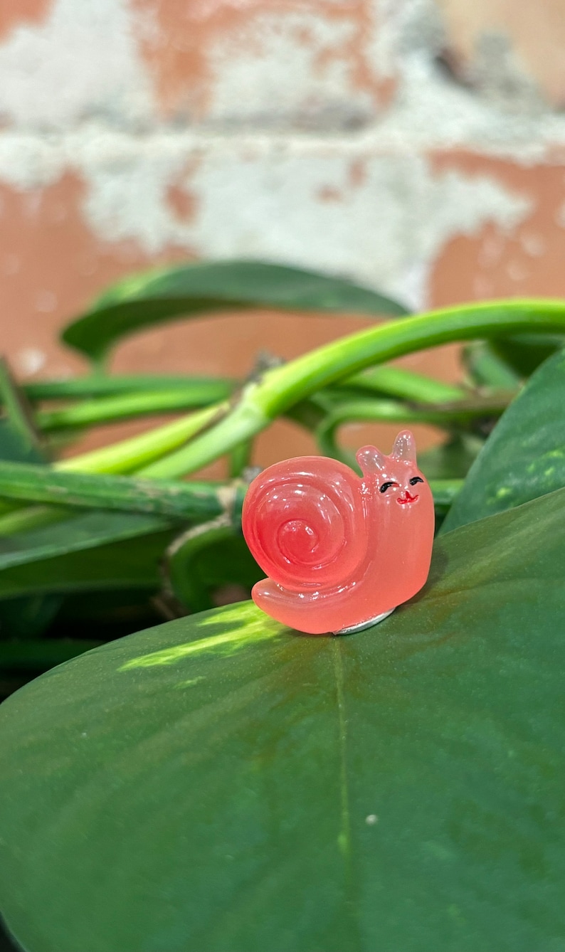 May include: A translucent, coral-colored snail figurine with a smiling face and black eyes. The snail is positioned on a large, green leaf, with a blurred background of more green leaves and a brick wall.