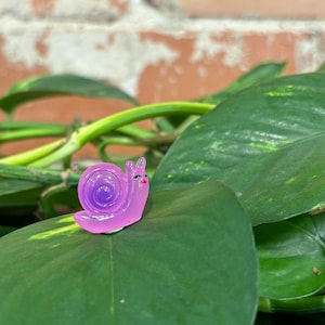May include: A small, whimsical, lavender-colored snail figurine with a spiraled shell and smiling face. The snail is perched on a large, green leaf, with a brick wall and green vines in the background. The figurine has a glossy finish.