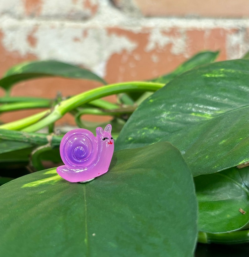 May include: A small, whimsical, lavender-colored snail figurine with a spiraled shell and smiling face. The snail is perched on a large, green leaf, with a brick wall and green vines in the background. The figurine has a glossy finish.