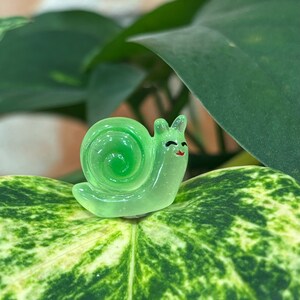 May include: A small, translucent green snail figurine with a smiling face and a spiral shell. The snail is resting on a large, green and white patterned leaf, with a blurred background of other leaves and a brick wall.