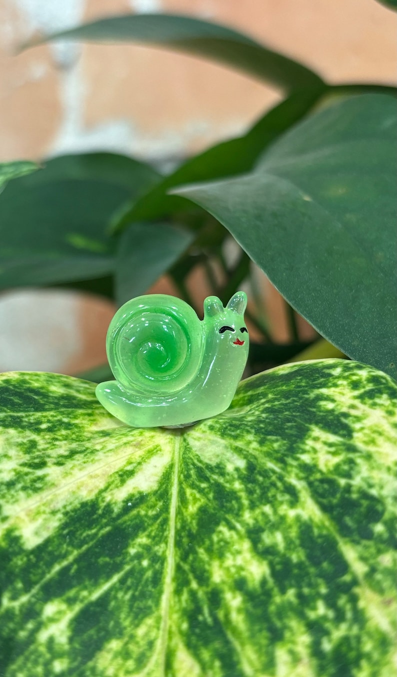 May include: A small, translucent green snail figurine with a smiling face and a spiral shell. The snail is resting on a large, green and white patterned leaf, with a blurred background of other leaves and a brick wall.