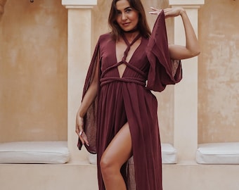 May include: A burgundy maxi dress with a high slit and a plunging neckline. The dress features a braided rope detail at the waist and a choker-style neckline. The sleeves are long and flowing. The background is a neutral-toned wall with decorative lanterns.