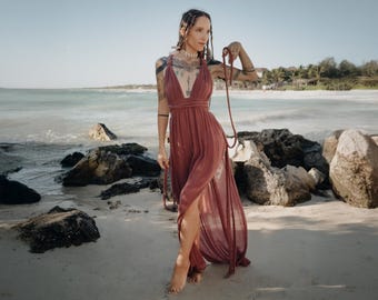 May include: A flowing, dusty rose maxi dress with a deep V-neck and high slit. The dress is worn on a beach with a clear blue sky and ocean in the background. A long, beaded necklace is held in the hand.