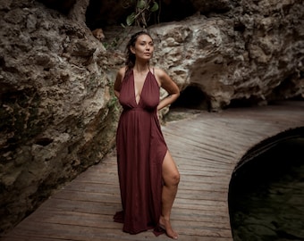 May include: A burgundy halter maxi dress with a deep V-neck and a high slit. The dress is worn outdoors on a wooden walkway, with a natural rock background.