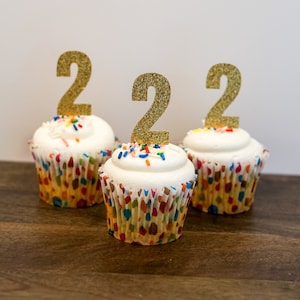 May include: Three white cupcakes with white frosting and colorful sprinkles. Each cupcake has a gold glitter number "2" topper.