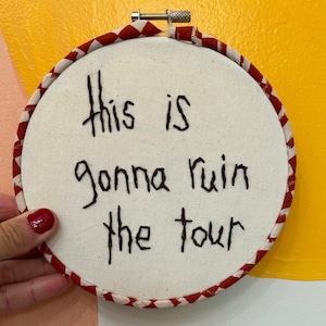 Może przedstawiać: Białe tamborek do haftu z czerwoną i białą kratą wokół krawędzi. Tamborek zawiera napis "This is gonna ruin the tour" wyszyty czarną nicią.