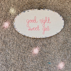 Może przedstawiać: Biały, owalny tamborek do haftu z kwiatowym wzorem wokół krawędzi. Tamborek ma haftowany napis "dobranoc słodka dziewczynko" w kolorze różowym.