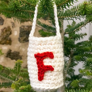 May include: White crocheted Christmas ornament with a red felt letter F hanging on a green Christmas tree.