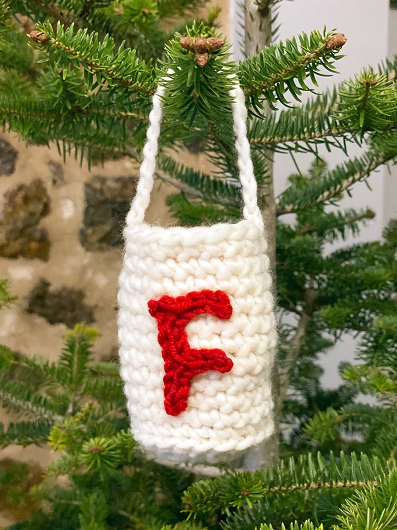 May include: White crocheted Christmas ornament with a red felt letter F hanging on a green Christmas tree.