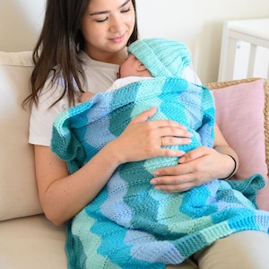 May include: A baby wrapped in a blue and turquoise knitted blanket and matching hat. The blanket features a geometric pattern. The baby is held by a person wearing a white shirt.