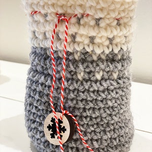 May include: A gray crocheted drawstring bag with a white crocheted top and a red and white twine tie. A wooden tag with a snowflake design is attached to the twine.