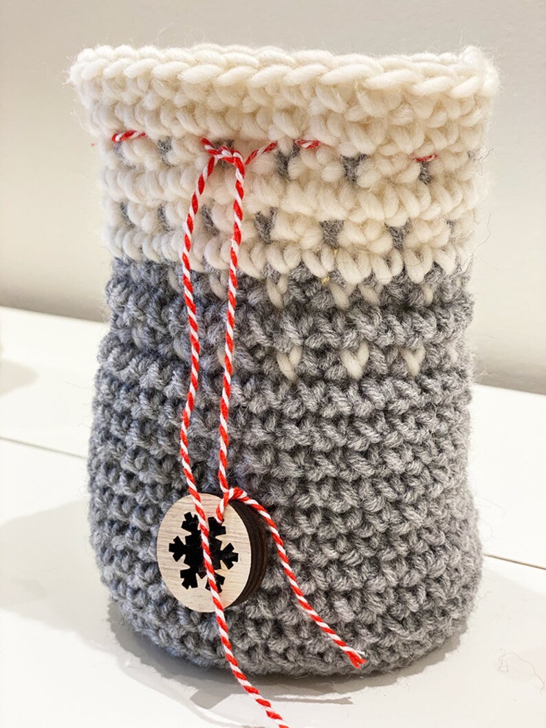 May include: A gray crocheted drawstring bag with a white crocheted top and a red and white twine tie. A wooden tag with a snowflake design is attached to the twine.