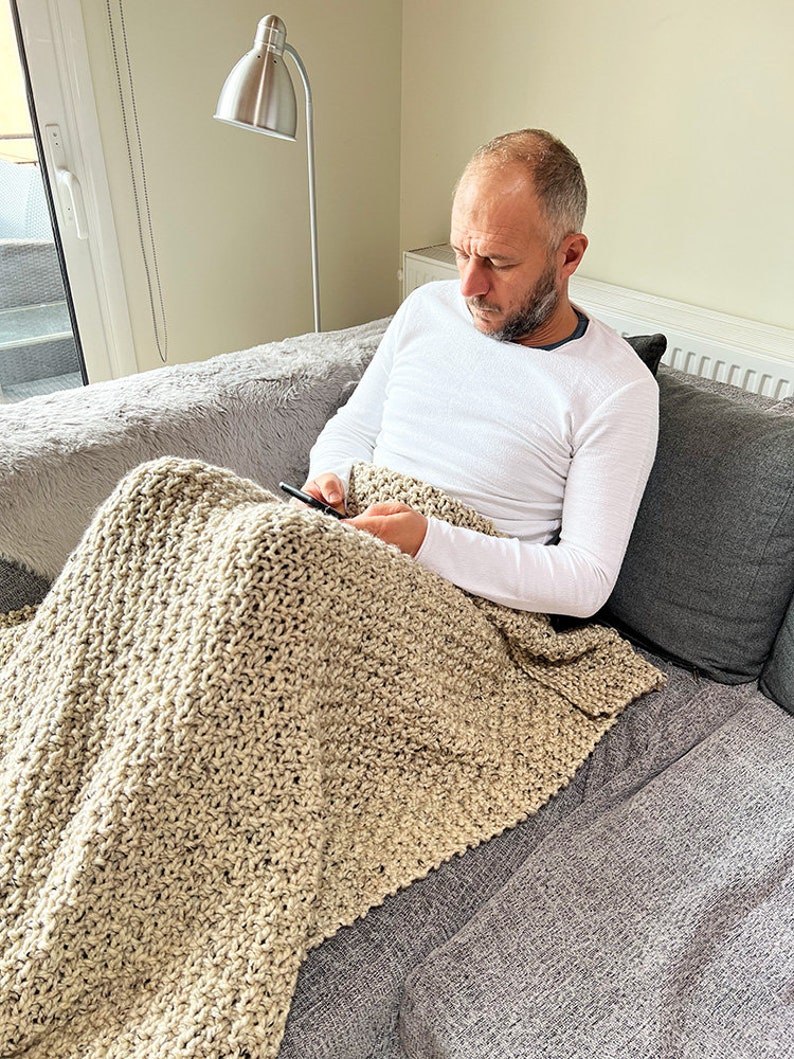 May include: A person sitting on a gray couch, wrapped in a beige knitted blanket, using a smartphone.