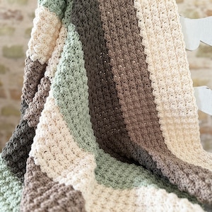 May include: A handmade crochet blanket in a striped pattern. The blanket features alternating stripes of cream, sage green, and various shades of brown. The textured stitch creates a cozy, inviting look. The blanket is draped over a white surface.