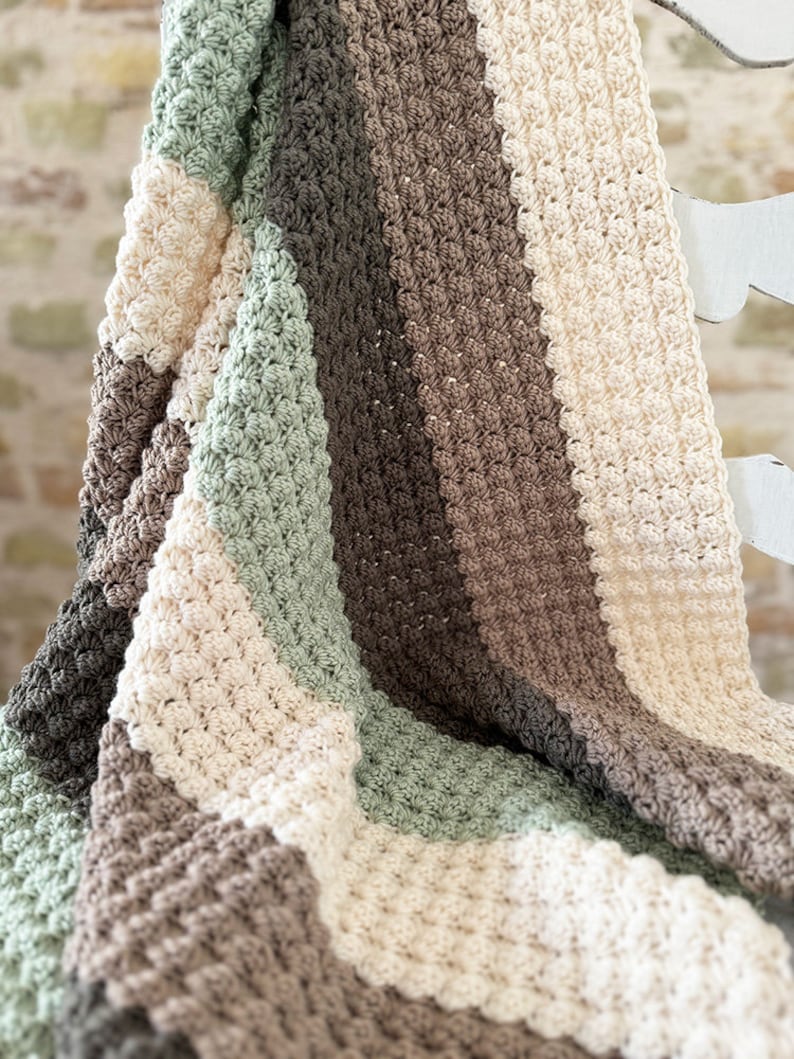 May include: A handmade crochet blanket in a striped pattern. The blanket features alternating stripes of cream, sage green, and various shades of brown. The textured stitch creates a cozy, inviting look. The blanket is draped over a white surface.