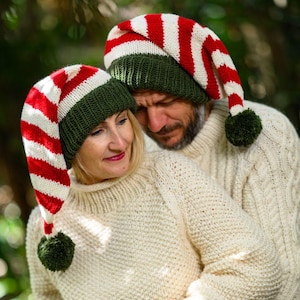 May include: Two people wearing matching Christmas elf hats. The hats are red and white striped with a green band and a green pom-pom. They are also wearing cream-colored, knitted sweaters. The background is a blurred green.