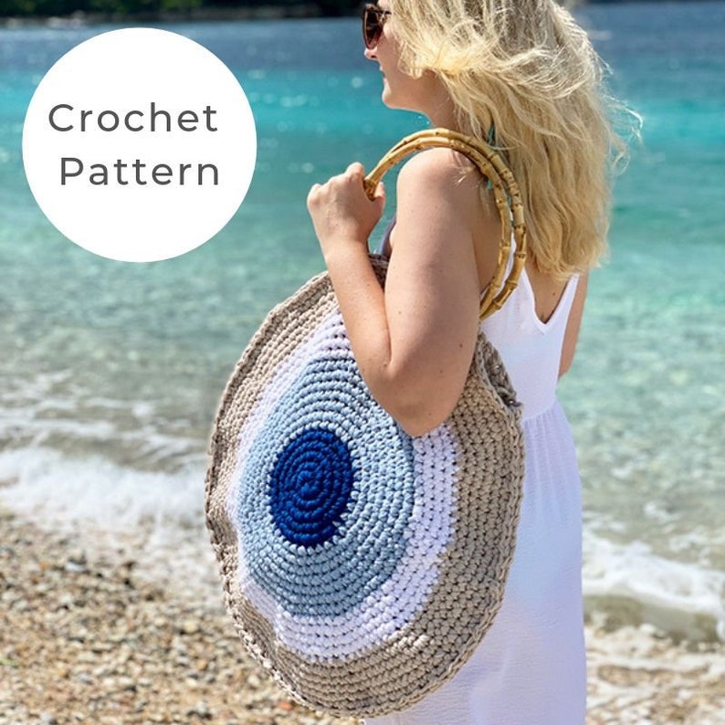 May include: A round, crocheted beach bag with a blue, white, and tan color scheme. The bag has bamboo handles and is being held by a person on a beach. The text "Crochet Pattern" is in a white circle in the upper left corner.