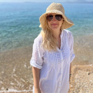 Könnte beinhalten: Eine Frau in einem weißen Hemd mit Knöpfen, einem Strohhut und einer Sonnenbrille steht am Strand.