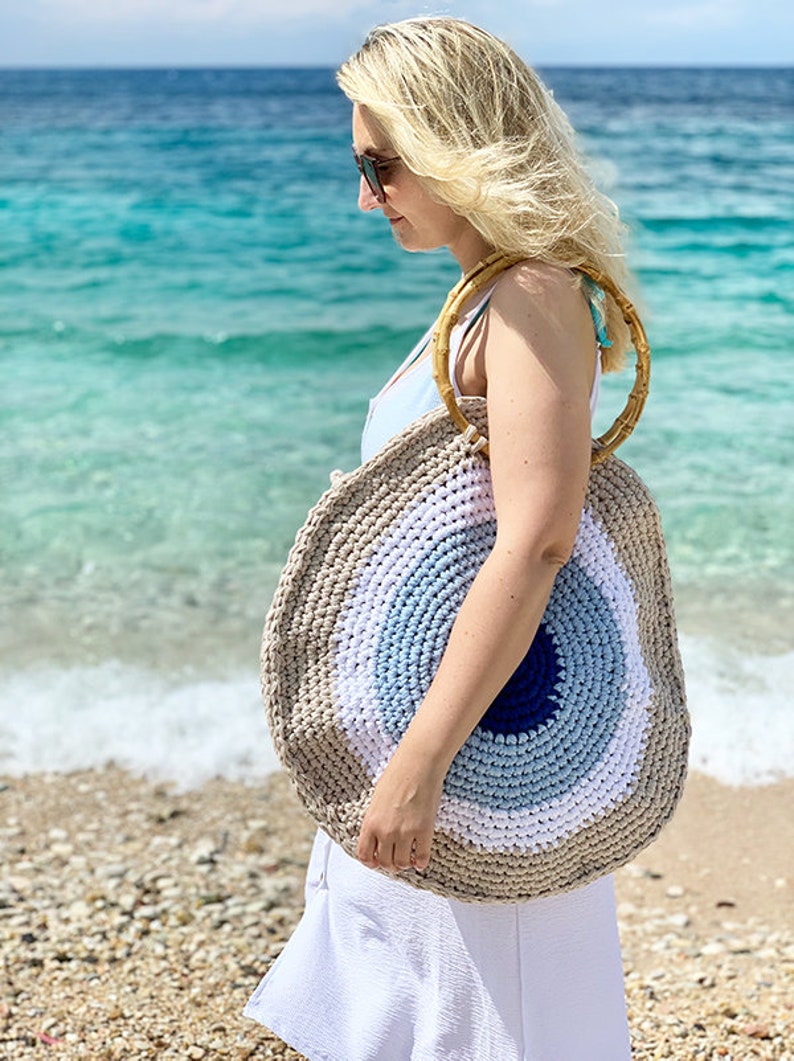 May include: A round, crocheted beach bag with a large eye design. The bag features concentric circles of beige, white, light blue, and dark blue. It has bamboo handles and is carried on the shoulder.