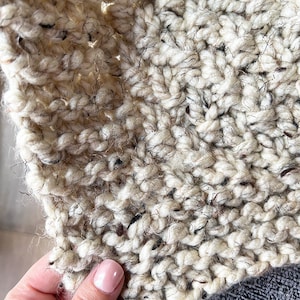 May include: Close-up of a cream-colored knitted fabric with a speckled texture. The fabric is soft and fluffy, with a slightly fuzzy appearance.