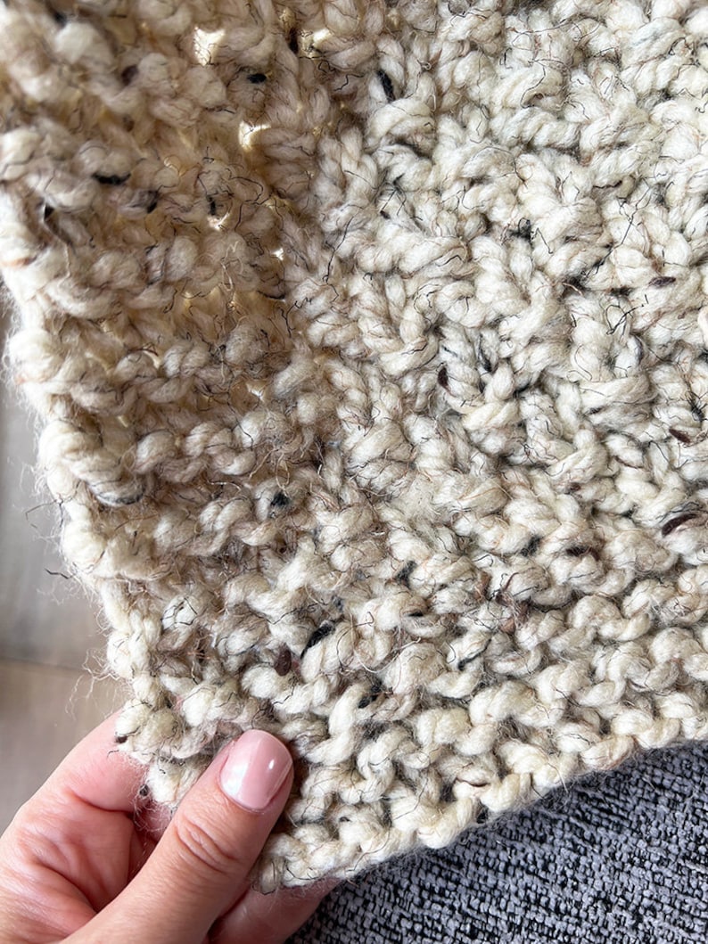 May include: Close-up of a cream-colored knitted fabric with a speckled texture. The fabric is soft and fluffy, with a slightly fuzzy appearance.