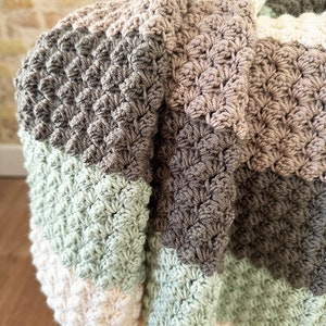 May include: A handmade crochet blanket in a neutral color palette. The blanket features a textured stitch pattern and is composed of stripes in shades of cream, sage green, gray, and brown. The blanket is draped over a white surface.