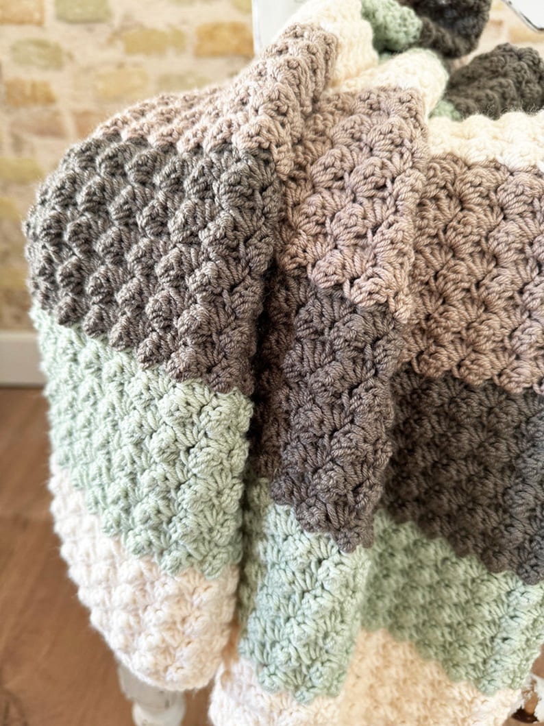 May include: A handmade crochet blanket in a neutral color palette. The blanket features a textured stitch pattern and is composed of stripes in shades of cream, sage green, gray, and brown. The blanket is draped over a white surface.