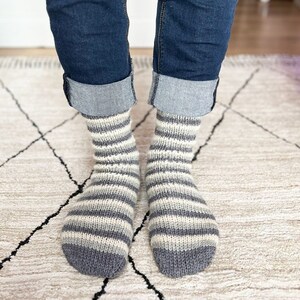 Könnte beinhalten: Ein Paar gestreifte Socken in Grau und Weiß. Die Socken sind aus weichem, gestricktem Stoff und haben eine bequeme Passform.