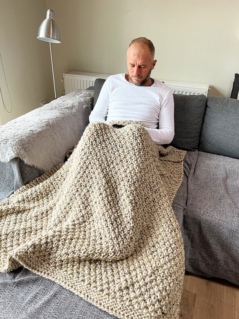 May include: A person sitting on a couch with a beige knitted throw blanket draped over their legs. The blanket has a textured, chunky knit pattern.