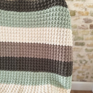 May include: A handmade, striped crochet blanket in shades of green, cream, brown, and gray. The blanket has a textured, bobble stitch pattern. The blanket is draped over a chair, showcasing the soft texture and color palette.