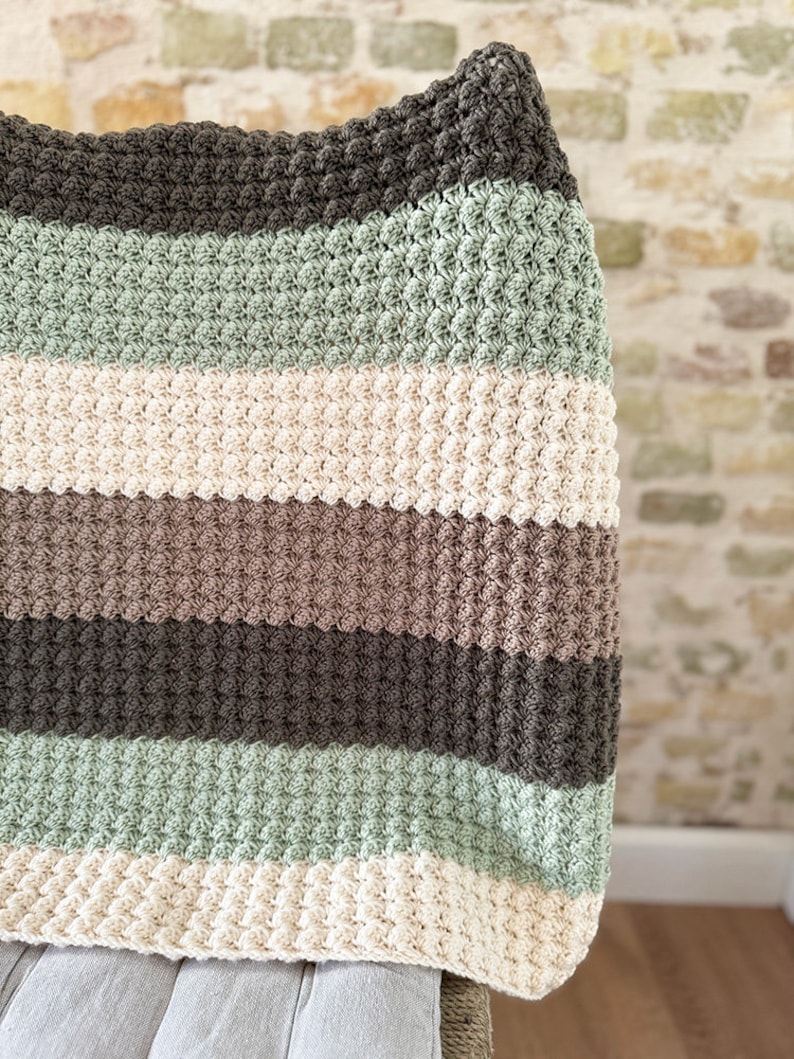 May include: A handmade, striped crochet blanket in shades of green, cream, brown, and gray. The blanket has a textured, bobble stitch pattern. The blanket is draped over a chair, showcasing the soft texture and color palette.