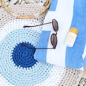 May include: A beach scene featuring a crocheted tote bag with a wooden handle, sunglasses, a striped blue and white towel, and a bottle of sunscreen. The bag has a spiral design in shades of blue and beige.