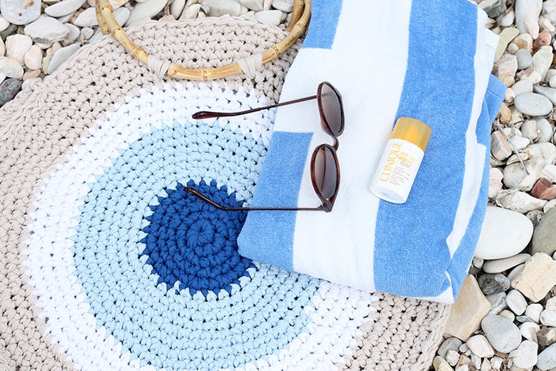 May include: A beach scene featuring a crocheted tote bag with a wooden handle, sunglasses, a striped blue and white towel, and a bottle of sunscreen. The bag has a spiral design in shades of blue and beige.