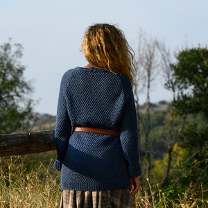 K&ouml;nnte beinhalten: Eine Frau tr&auml;gt einen blauen Strickpullover mit einem braunen G&uuml;rtel und steht auf einem Feld mit hohem Gras und B&auml;umen im Hintergrund.