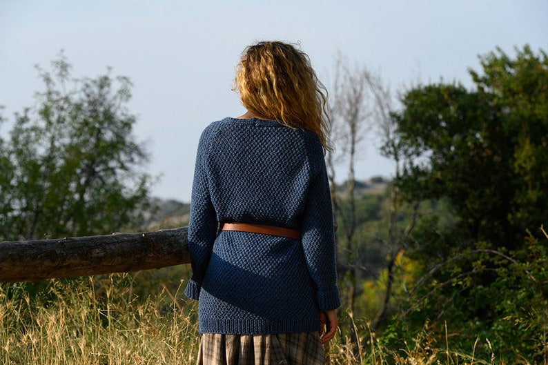 K&ouml;nnte beinhalten: Eine Frau tr&auml;gt einen blauen Strickpullover mit einem braunen G&uuml;rtel und steht auf einem Feld mit hohem Gras und B&auml;umen im Hintergrund.