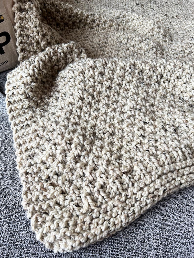 May include: A close-up of a beige knitted throw blanket with a textured pattern. The blanket is made of a soft, fluffy yarn and has a warm, cozy feel.