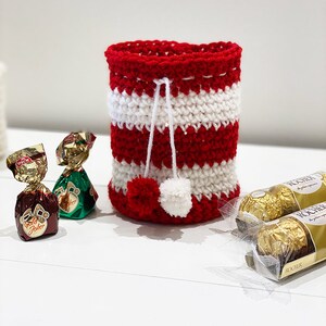 May include: Red and white striped crocheted gift bag with a white pom-pom on top. The bag is filled with chocolate candies.