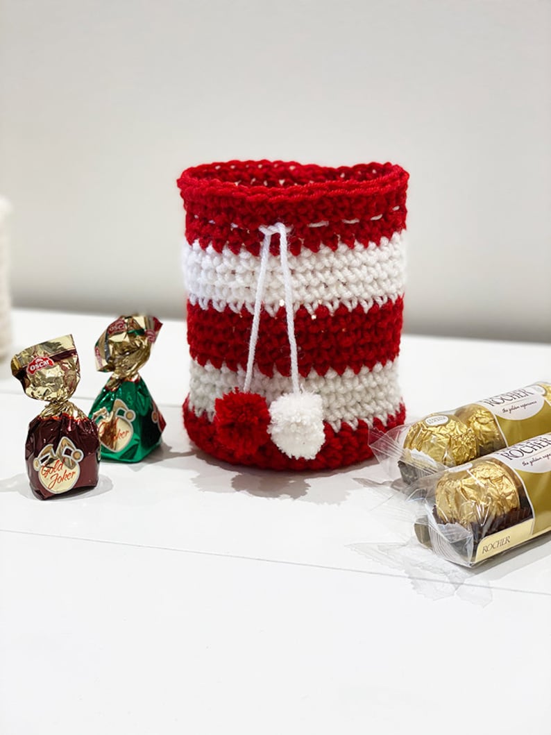 May include: Red and white striped crocheted gift bag with a white pom-pom on top. The bag is filled with chocolate candies.