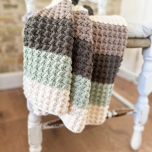 Puede incluir: Una manta texturizada hecha a mano, sobre una silla blanca. La manta presenta rayas horizontales en crema, verde salvia, marrón y gris. El punto de ganchillo crea un patrón en relieve, similar a bolitas. La silla tiene un aspecto vintage.