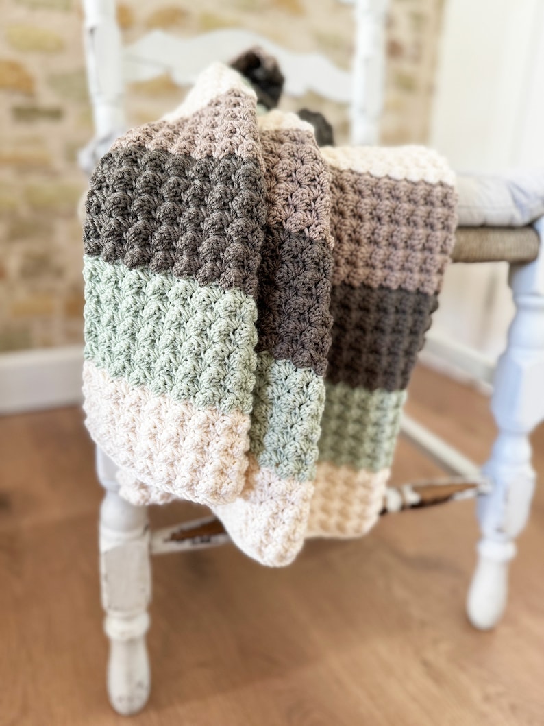 May include: A handmade, textured blanket draped over a white chair. The blanket features horizontal stripes in cream, sage green, brown, and gray. The crochet stitch creates a raised, bobble-like pattern. The chair has a distressed, vintage look.